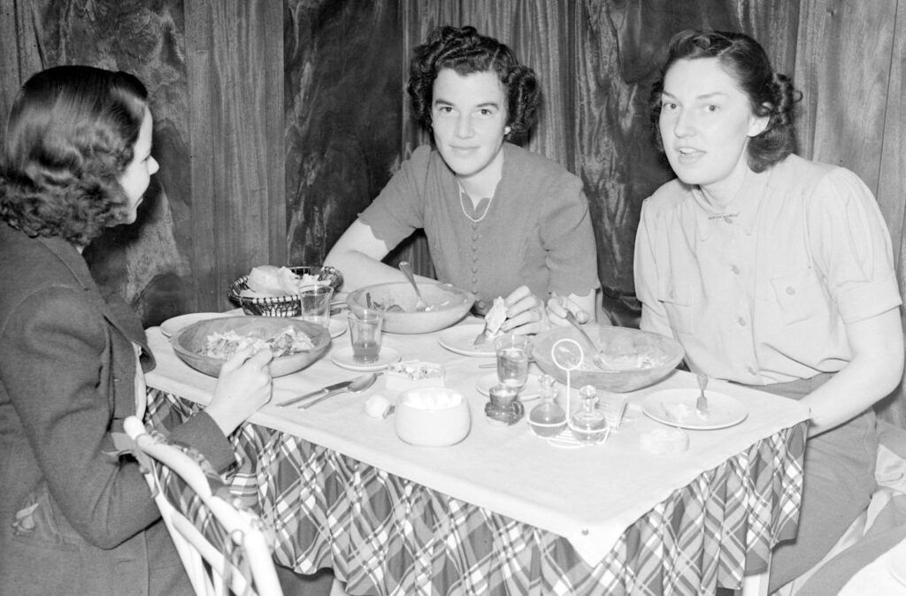 A black and white photograph of three women seated at a table in a restaurant eating salad in bowls