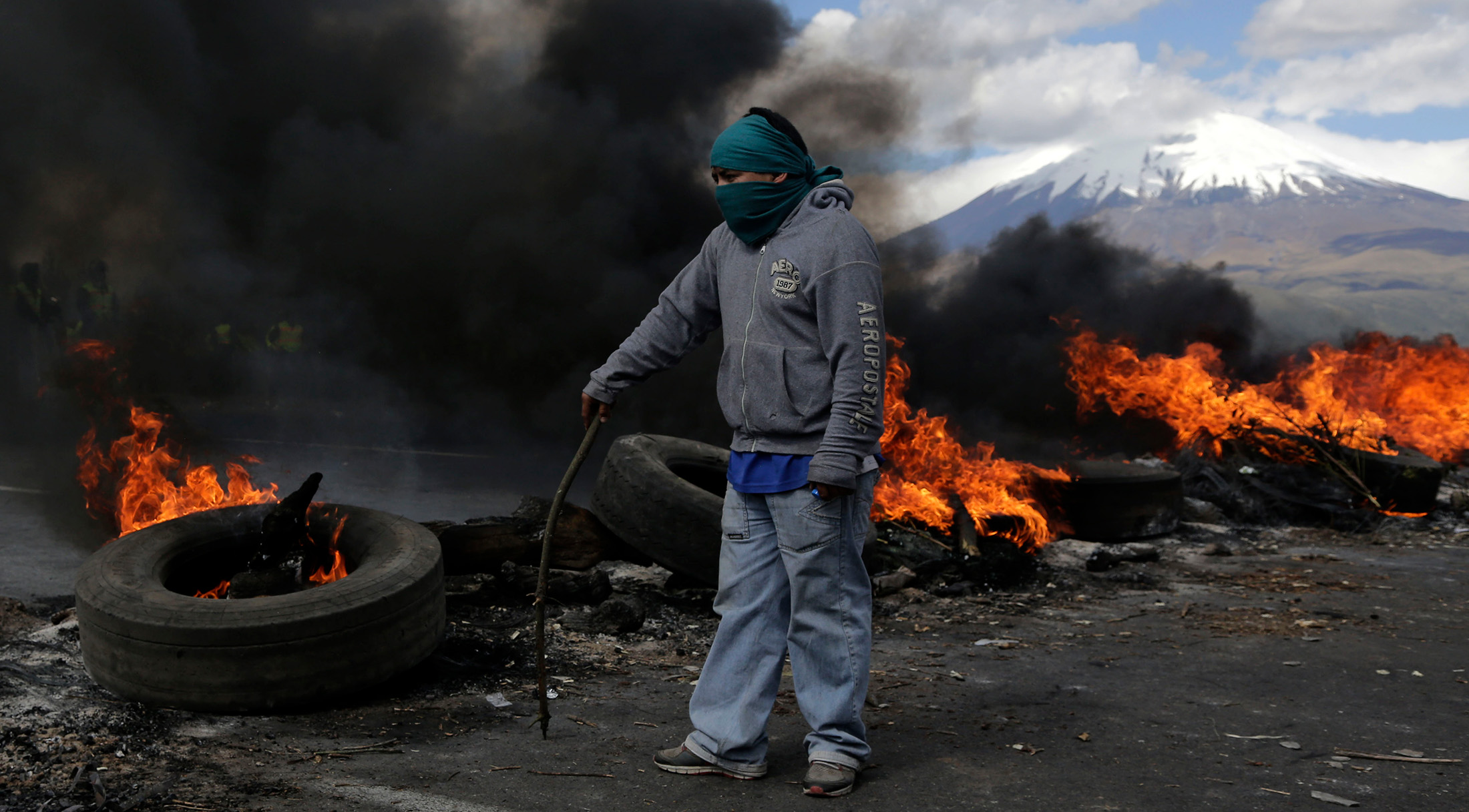 color photograph of a masked man standing in front of a roadblock of burning tires and trees, with a mountain in the background