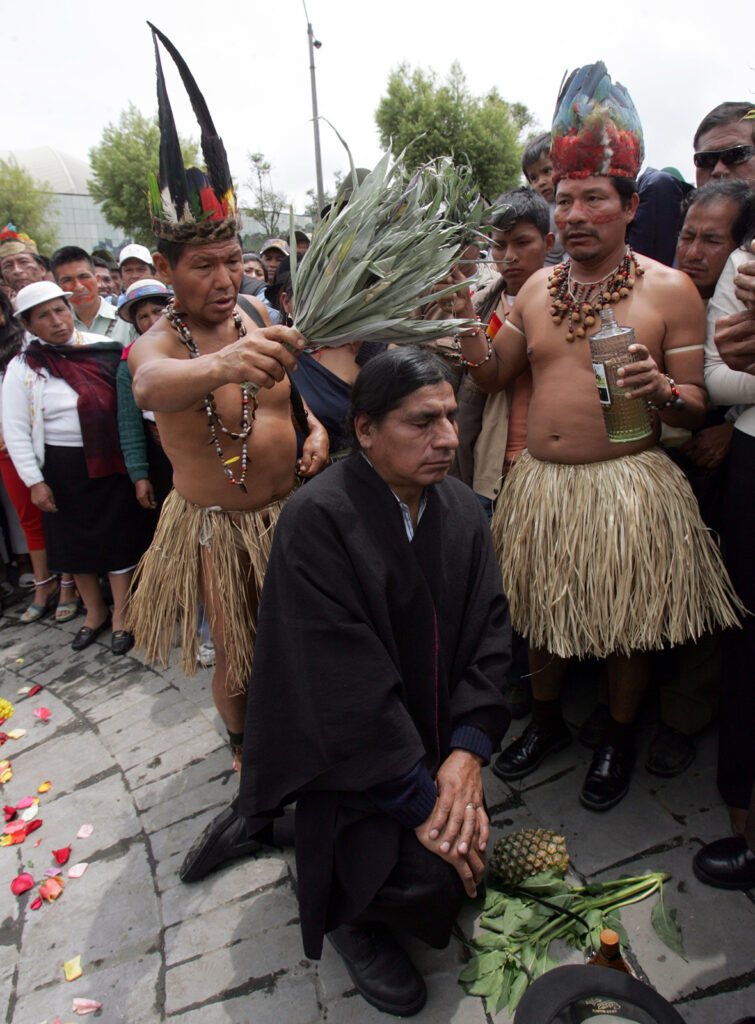 color photograph of a man kneeling in a crowd while two men in Indigenous dress hold plant leaves above his head