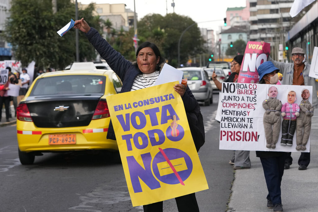 color photograph of a woman standing on a city sidewalk holding a sign that reads “Vota todo no.”