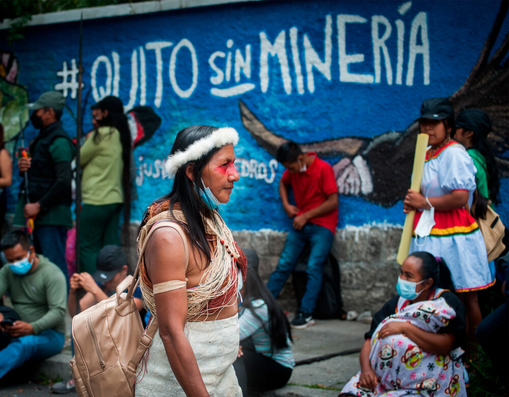 A color photo of a woman in a crowd walking past a painted blue wall with street art that reads: Quito sin minería (Quito without mining).