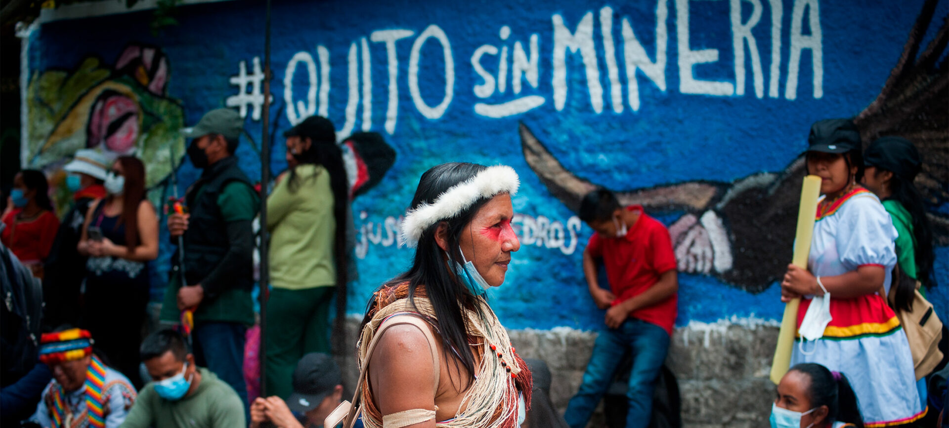 A color photo of a woman in a crowd walking past a painted blue wall with street art that reads: Quito sin minería (Quito without mining).