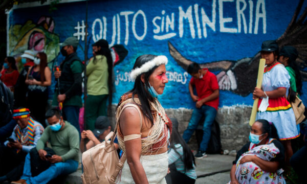 A color photo of a woman in a crowd walking past a painted blue wall with street art that reads: Quito sin minería (Quito without mining).