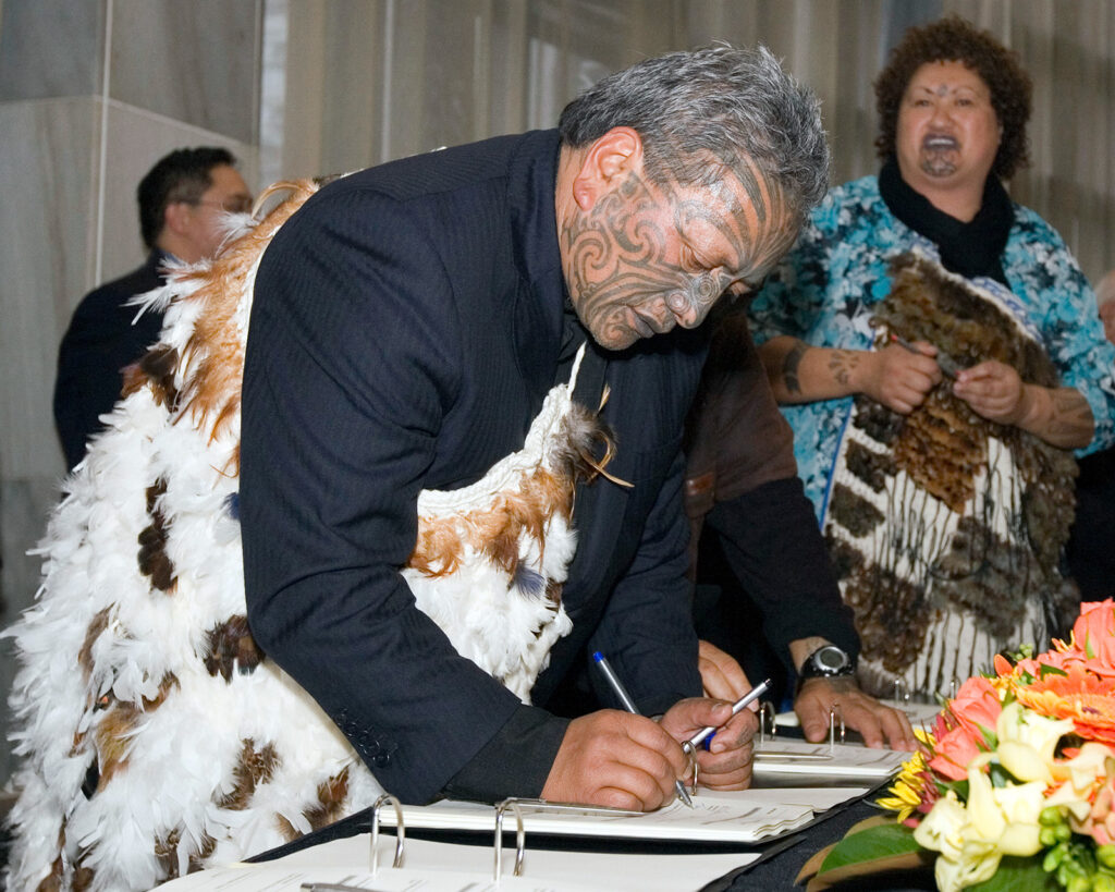 Color photo of a man with intricate face tattoos and ceremonial dress signing a document