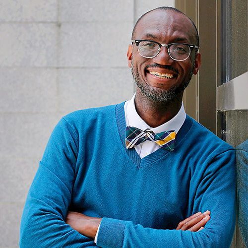 André Isaacs in blue sweater and bowtie leaning on a glass door