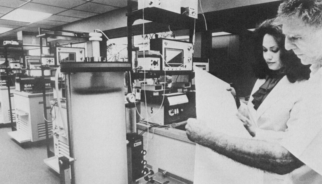 A man and woman reading from a large piece of paper in a lab filled with computers and scientific equipment