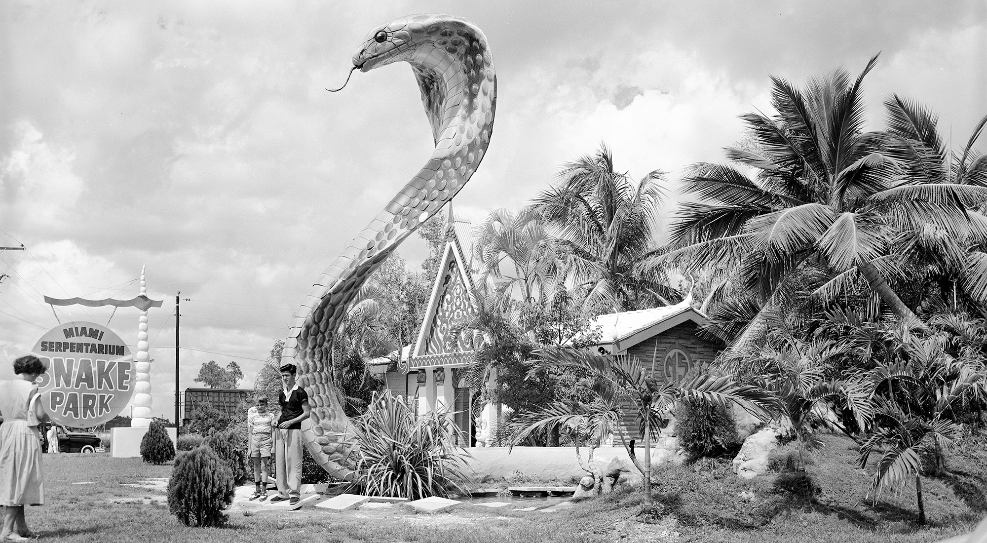 Two boys pose in front of an ornate building surrounded by tropical trees and plants with a large cobra statue in the front. A sign on the far left reads: “Miami Serpentarium Snake Park.”
