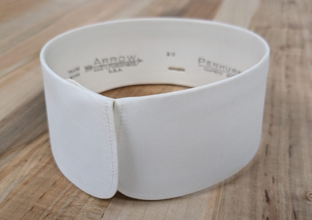A starched detachable white shirt collar curled into a circle. Inside the collar is black print that reads “Arrow USA.”
