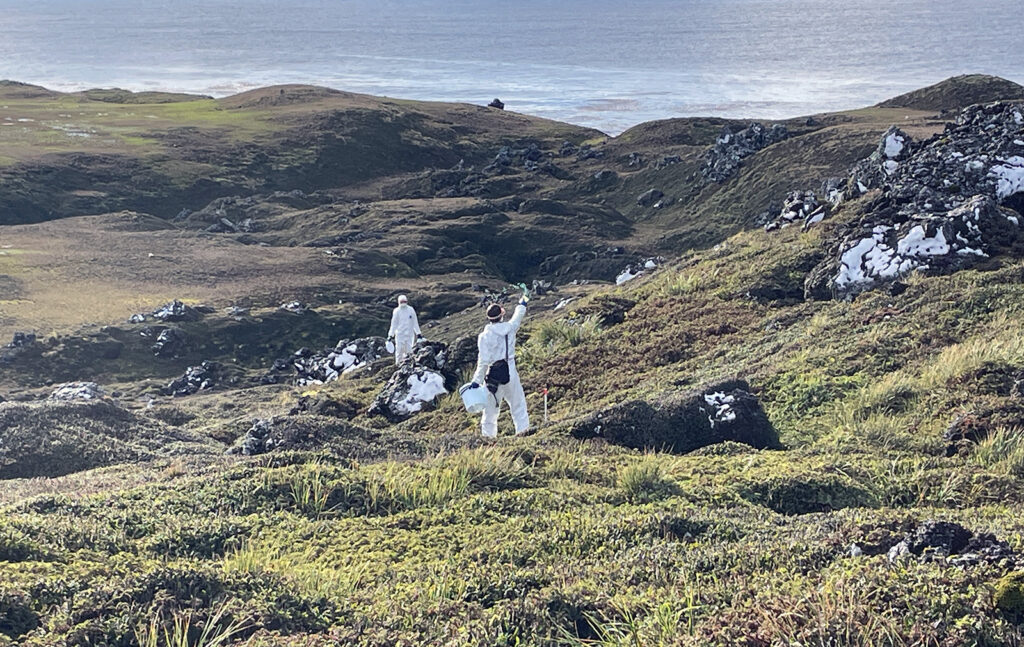 Two people in hazmat suits in a rocky, coastal landscape