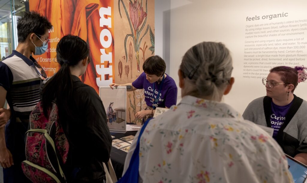 A woman stands behind a long table within a museum gallery, and she gestures to an image displayed on the table. Three individuals stand in front of the table facing the woman and are looking at the image.