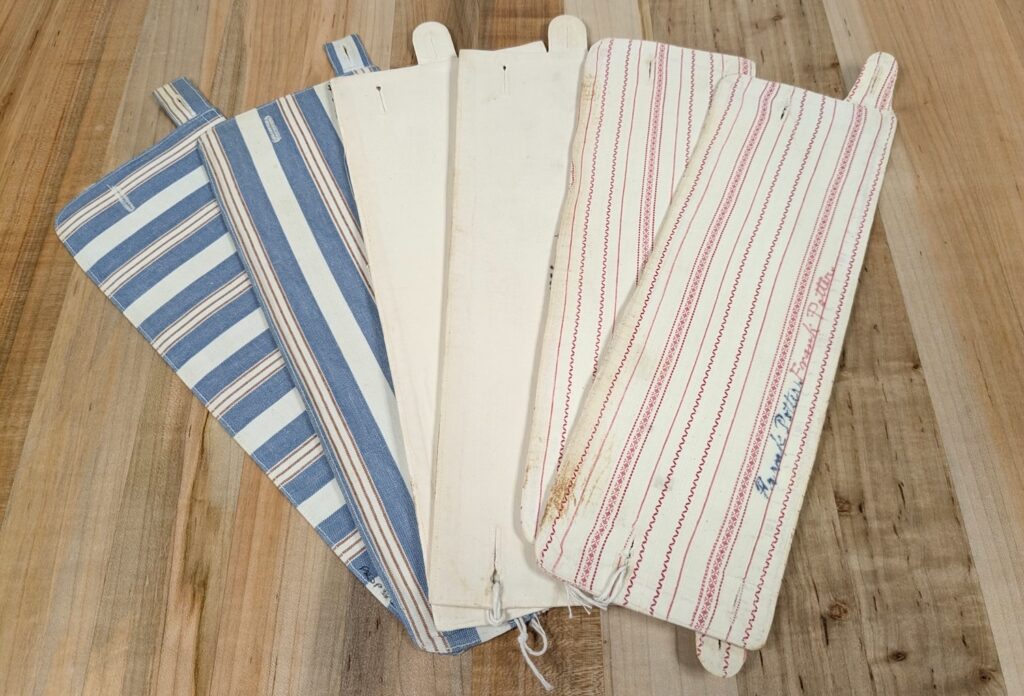 Three pairs of detachable shirt cuffs lying on a table. The shape of each flatten cuff is a long rectangle, and there are two buttonholes at each end. One pair has alternating blue, red, and white stripes, second pair is plain white, and third pair is white with thin red stripes.