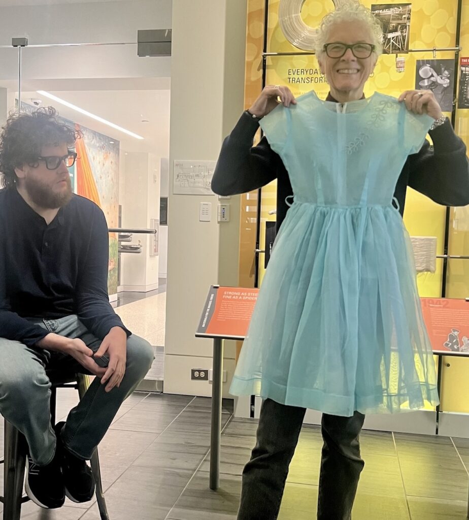A woman is holding up a child’s sheer blue dress. To her left, a man is seated on a stool. Behind them is a museum display on nylon fabric.
