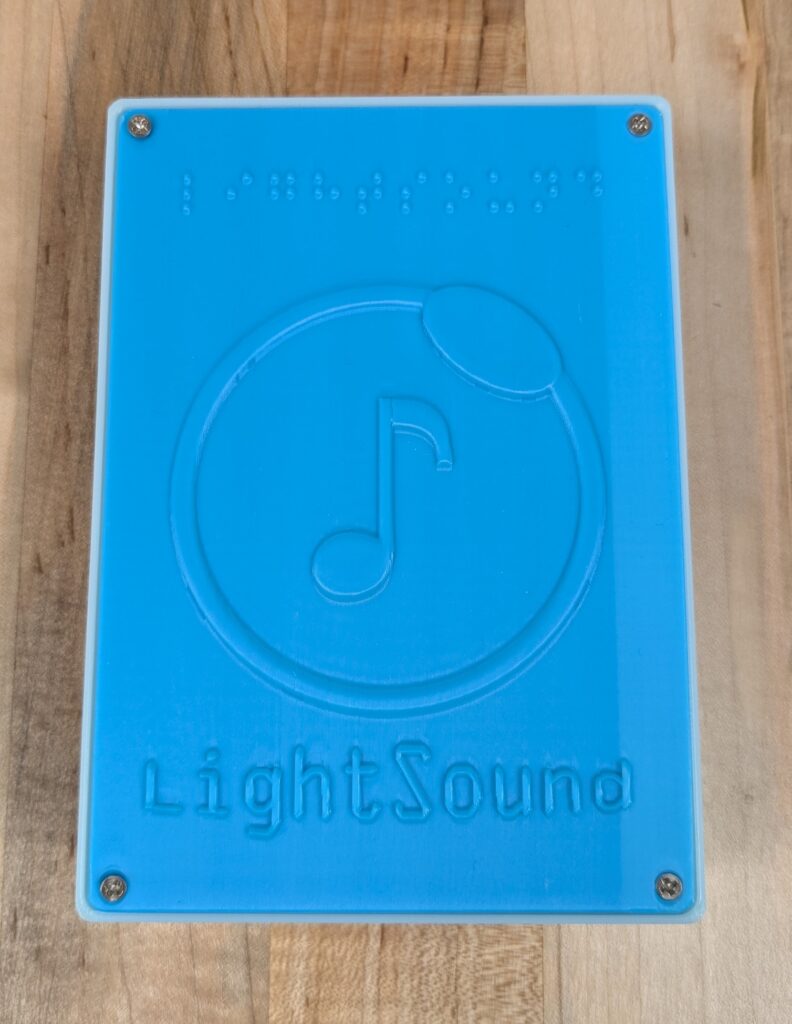 A blue rectangular plastic box with the word “LightSound” written in braille at the top. In the center is a tactile image of a music note surrounded by a circle. At the bottom is the word “LightSound” printed in raised text.