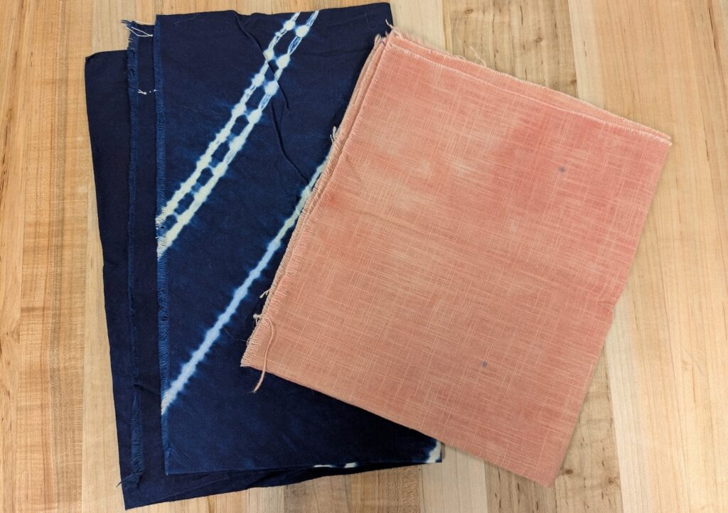 Two folded pieces of fabric. The piece of cloth on the left has been tie dyed; it is dark blue with white lines indicating where it was tied when dyed. The piece of cloth on the right is light red and faint lines can be seen indicating a woven pattern.