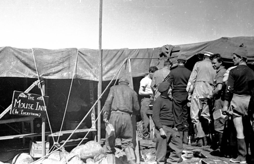 Men queuing in front of a tent with a sign reading “The Mouse Inn (In Everything)”