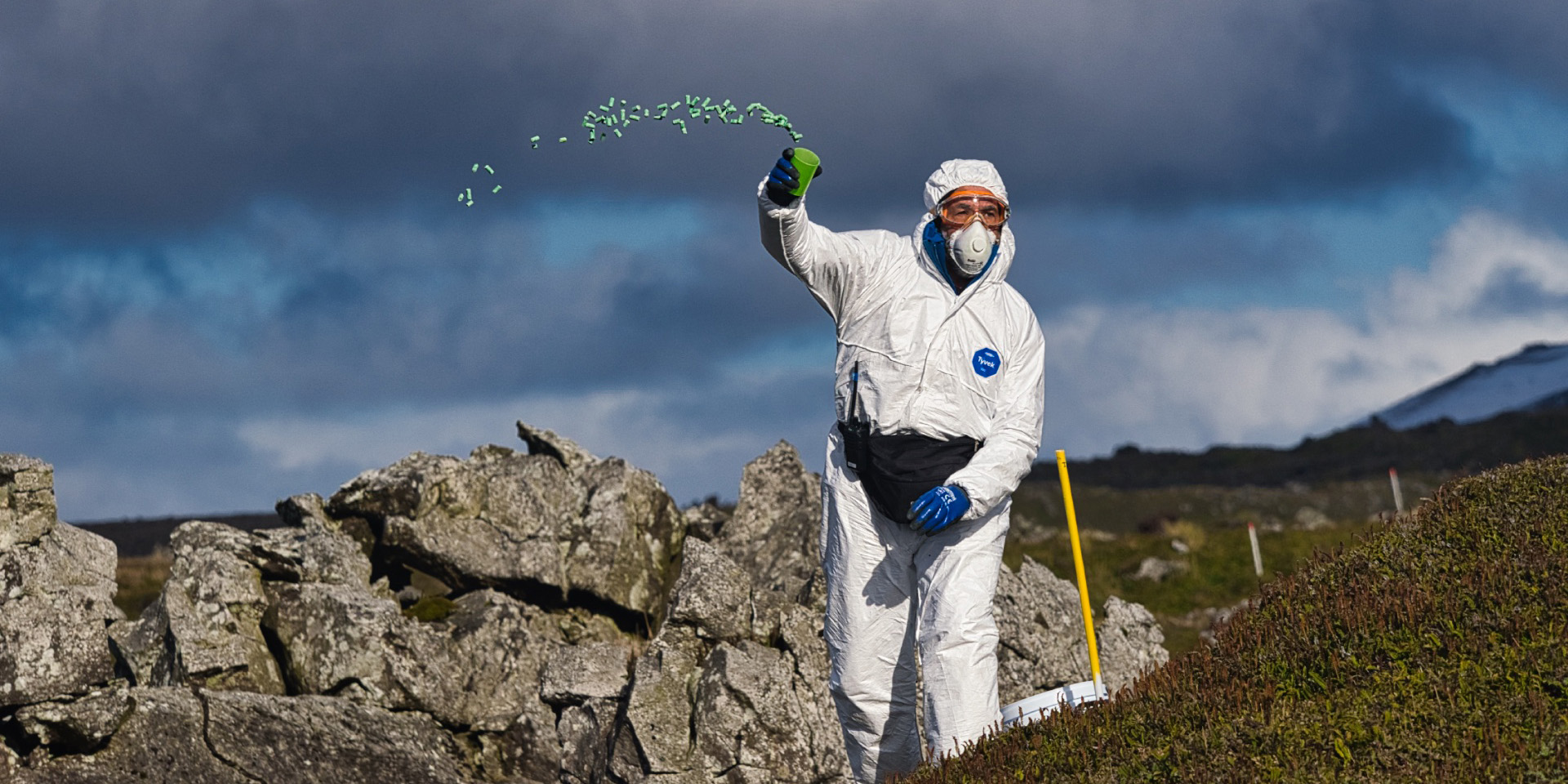Man in hazmat suit, goggles and face mask tossing green pellets