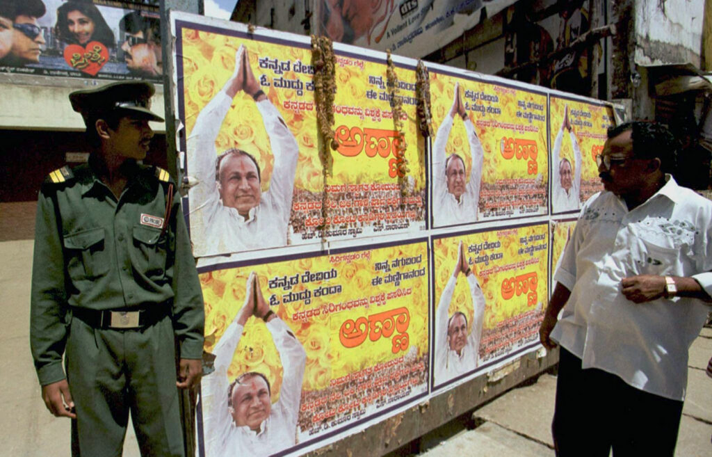 A man and a police officer observe posters featuring a man in white shirt with his arms raised and hands clasped in prayer.