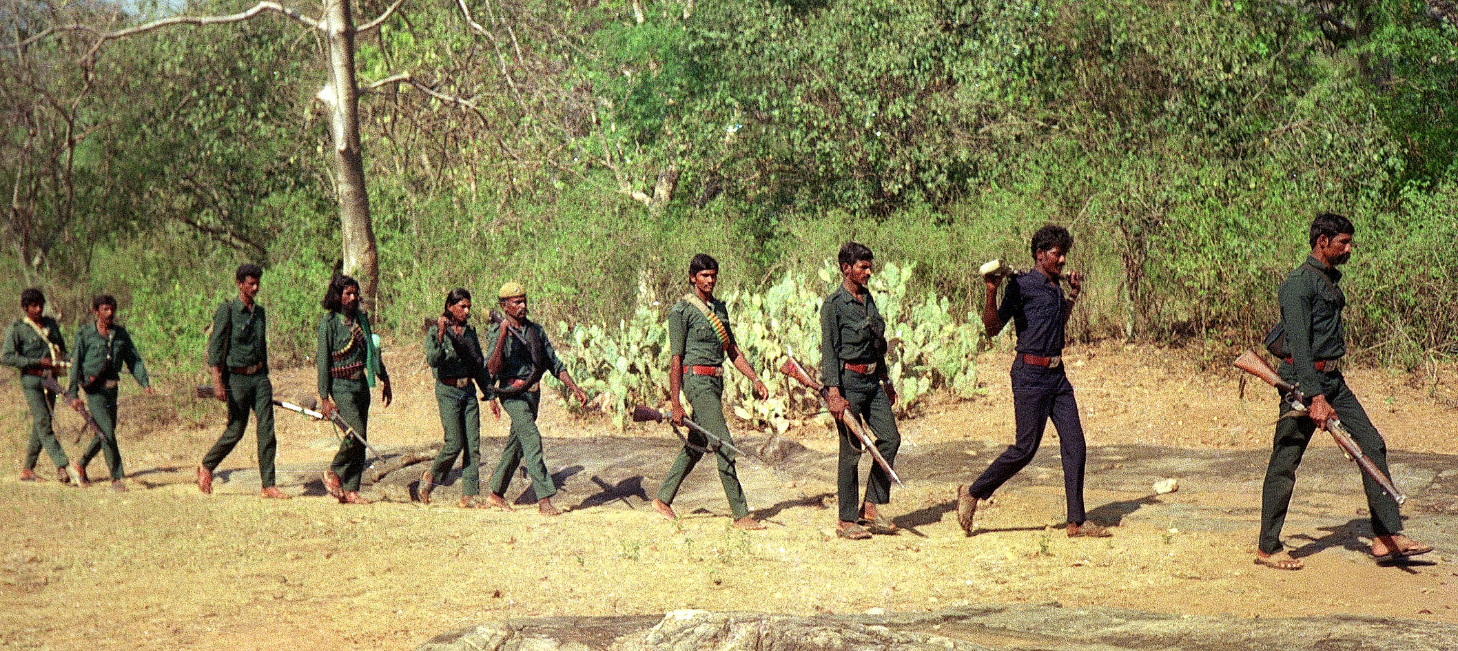 Line of ten armed men in green camo walking in single file