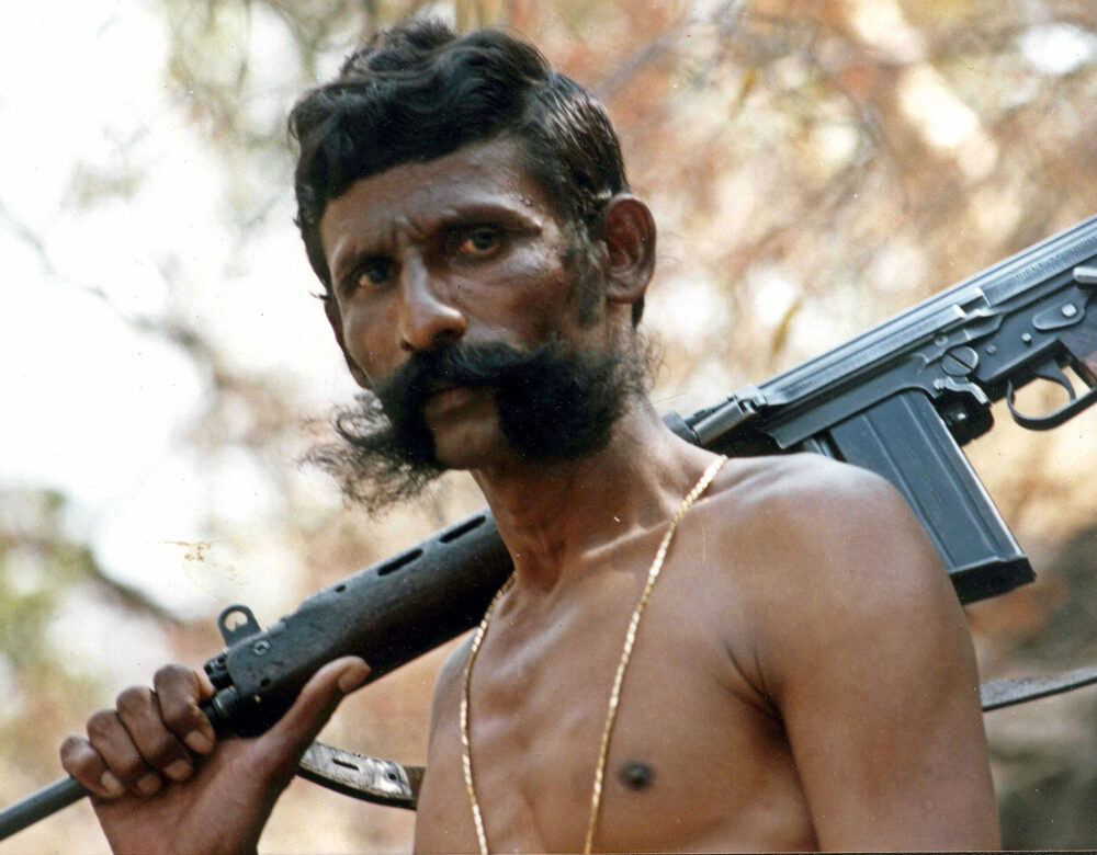 A shirtless man standing with a gun on his shoulder