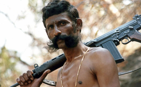 A shirtless man standing with a gun on his shoulder