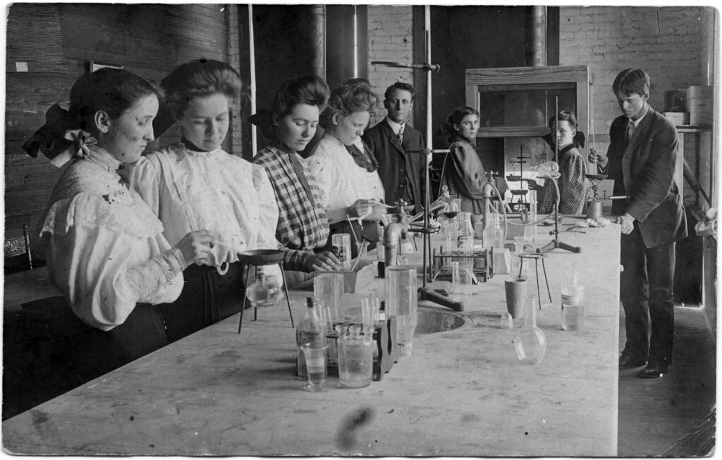 Six women and one man working at a lab bench with chemical instruments, while a male instructor observes.