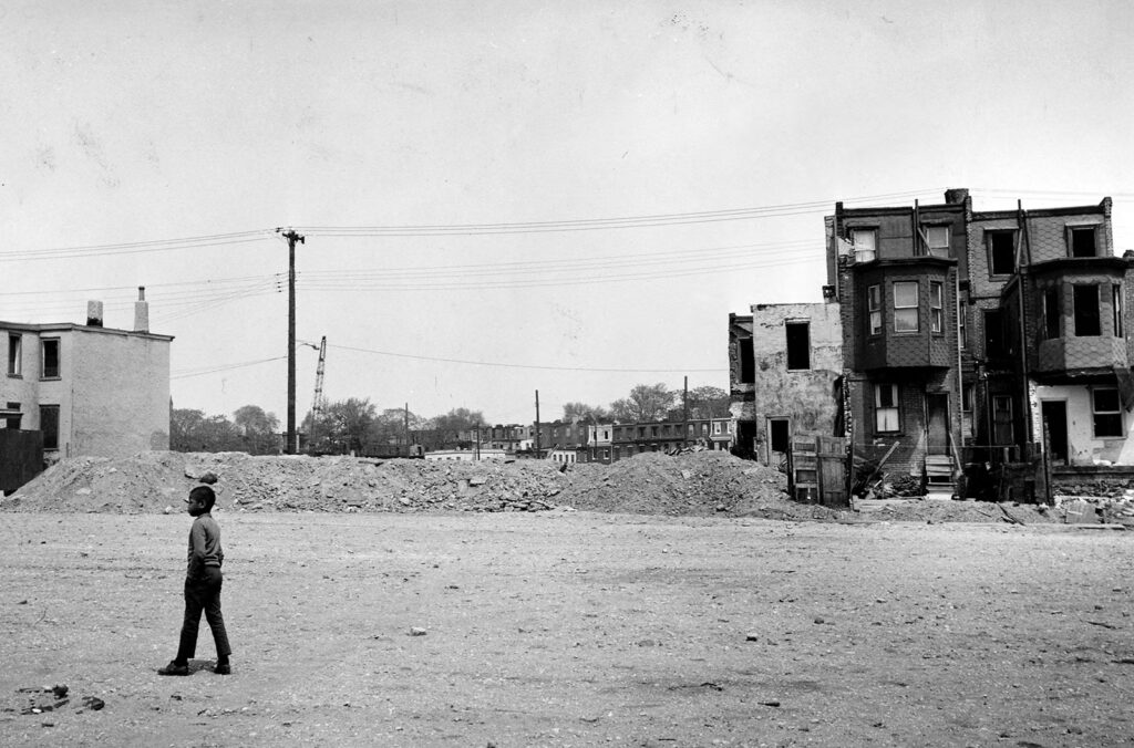 Boy standing in a leveled city lot with a few remaining houses in the background.