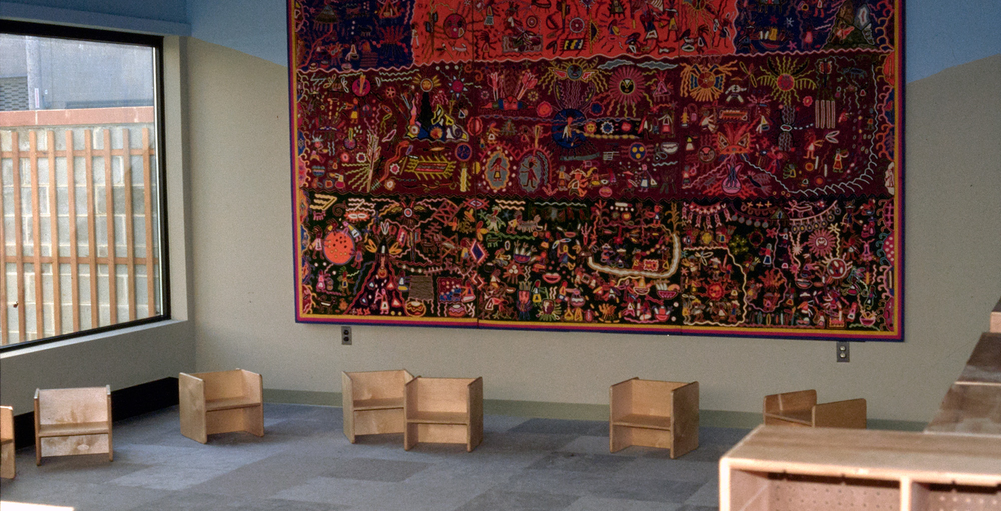 A large, colorful artwork hanging in a large room with small wooden chairs on the floor.