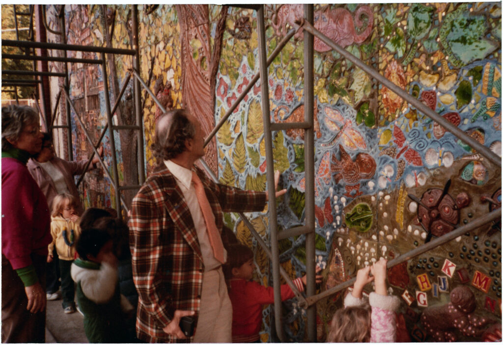 Man placing his hand on a colorful mural with scaffolding in front of it. Women and children are observing the mural around him.