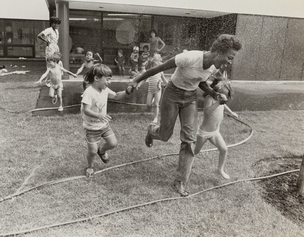 Women and children playing outdoors with a garden hose.