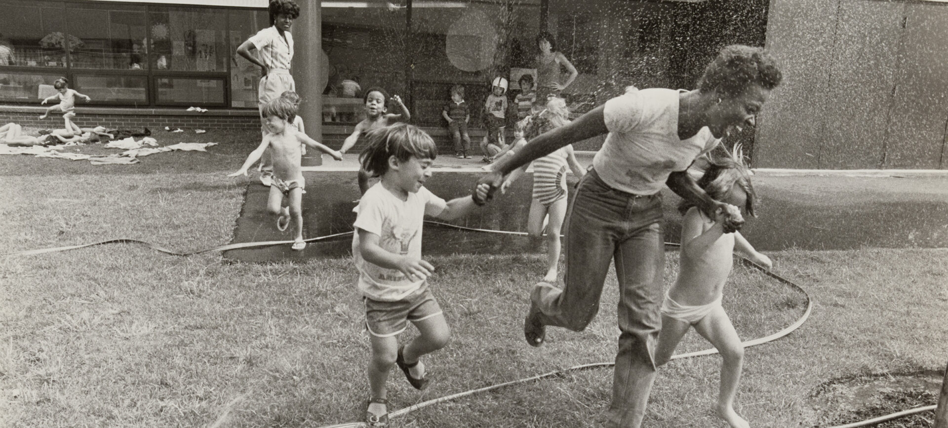 Women and children playing outdoors with a garden hose.