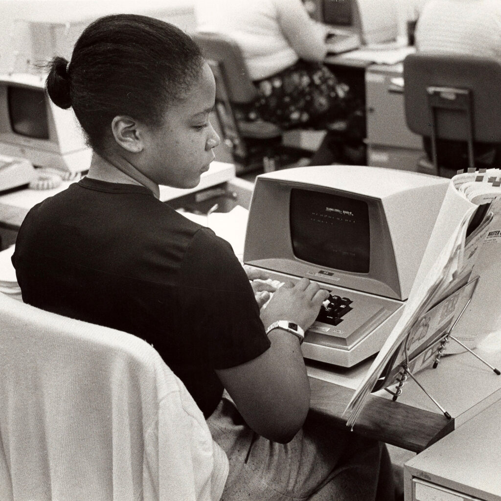 Woman seated at a desk in an office typing on an early computer terminal.