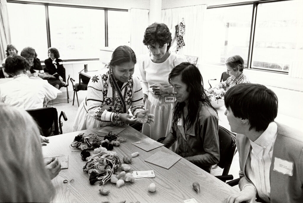 Group of people gathered around a table observing a woman with a piece of yarn in her hands.