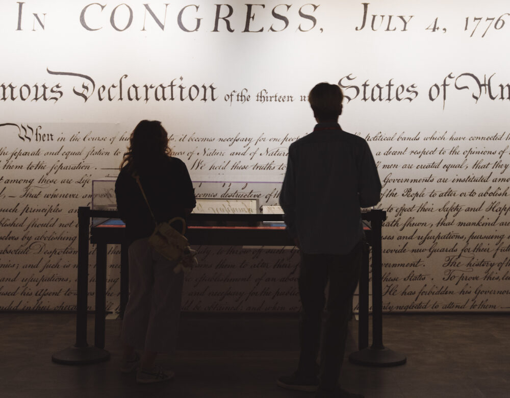 Two people in silhouette in front of a museum exhibit, a large detail of the Declaration of Independence blown up on the wall behind