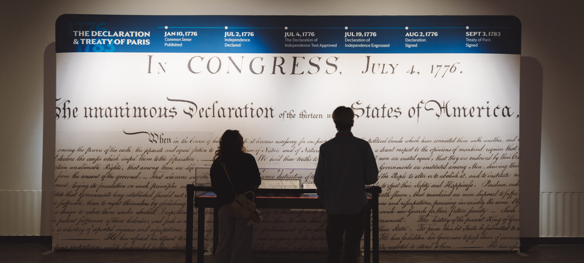Two people in silhouette in front of a museum exhibit, a large detail of the Declaration of Independence blown up on the wall behind