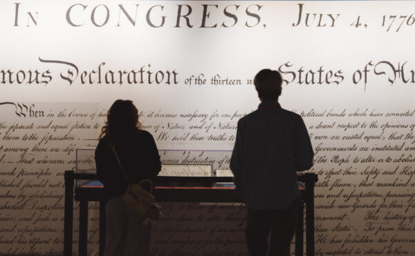Two people in silhouette in front of a museum exhibit, a large detail of the Declaration of Independence blown up on the wall behind