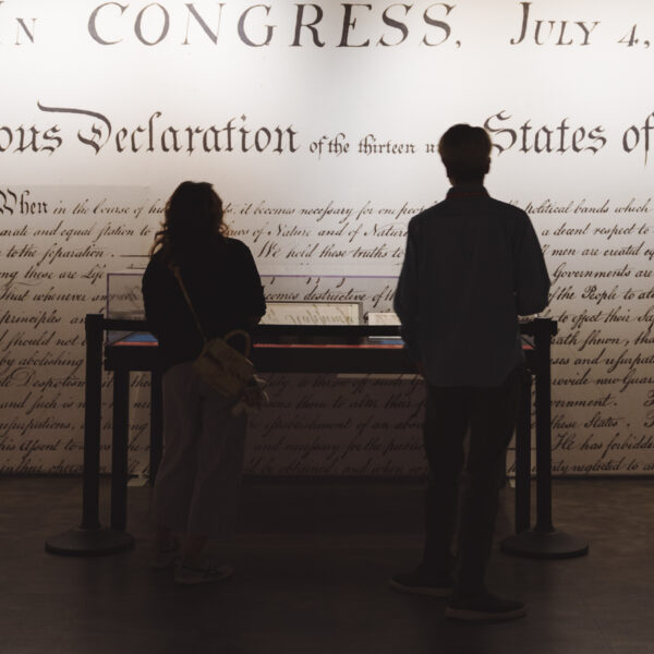 Two people in silhouette in front of a museum exhibit, a large detail of the Declaration of Independence blown up on the wall behind