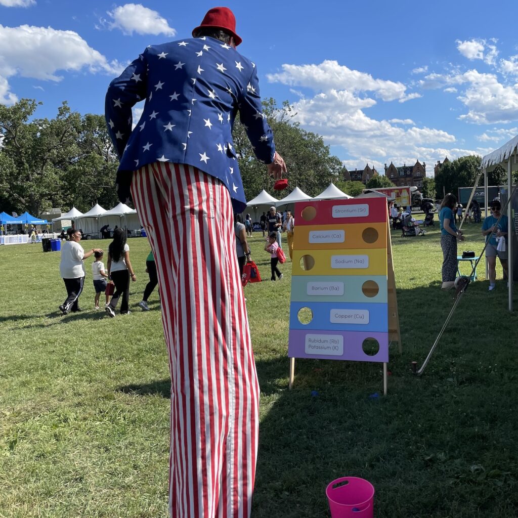 Man dressed in red, white and blue walking on stilts tosses a bean bag