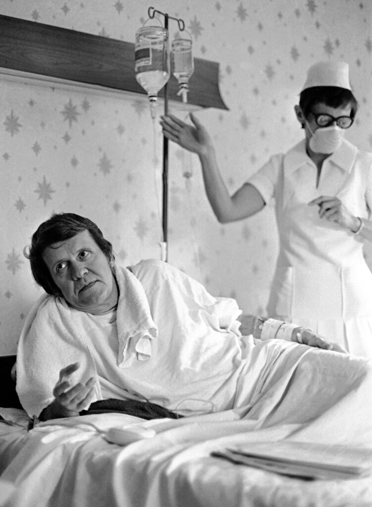 Man sitting up in a hospital bed while a nurse attends to his glass IV bottles.