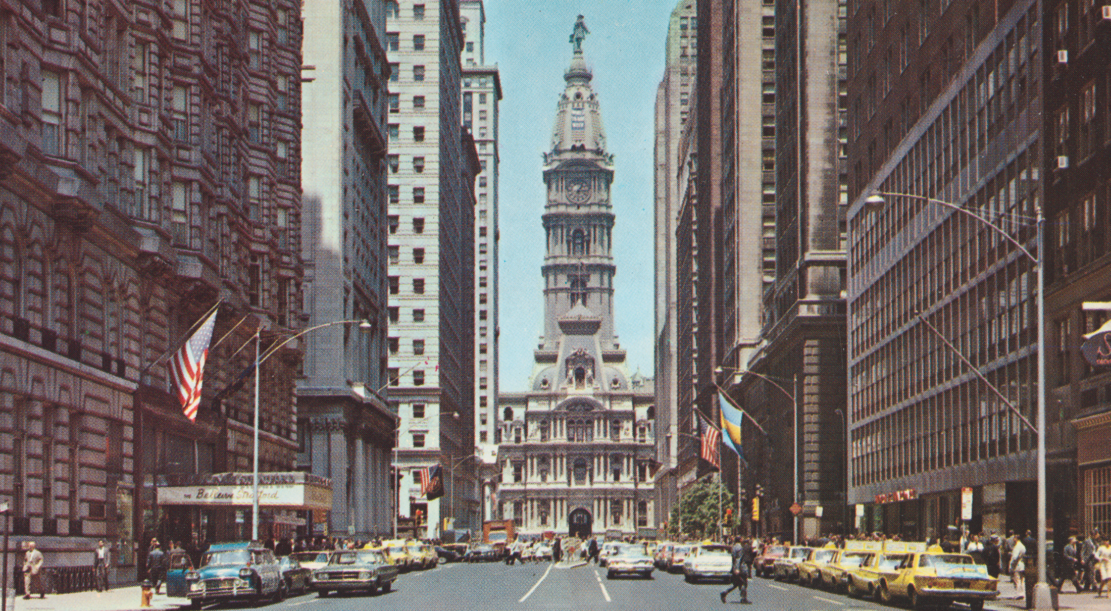 Cityscape with large buildings on each side. A large, ornate building with a statue of a man on the top is visible at the end of the street.