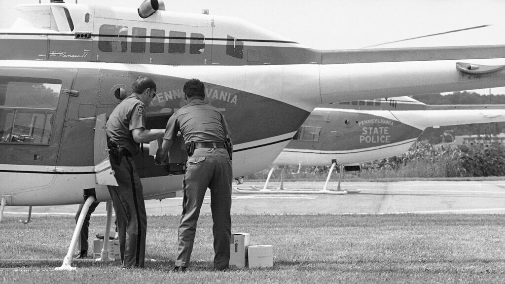 Two men loading boxes into a helicopter. A second helicopter is visible in the background. Text on the side of the helicopters reads: “Philadelphia State Police.”