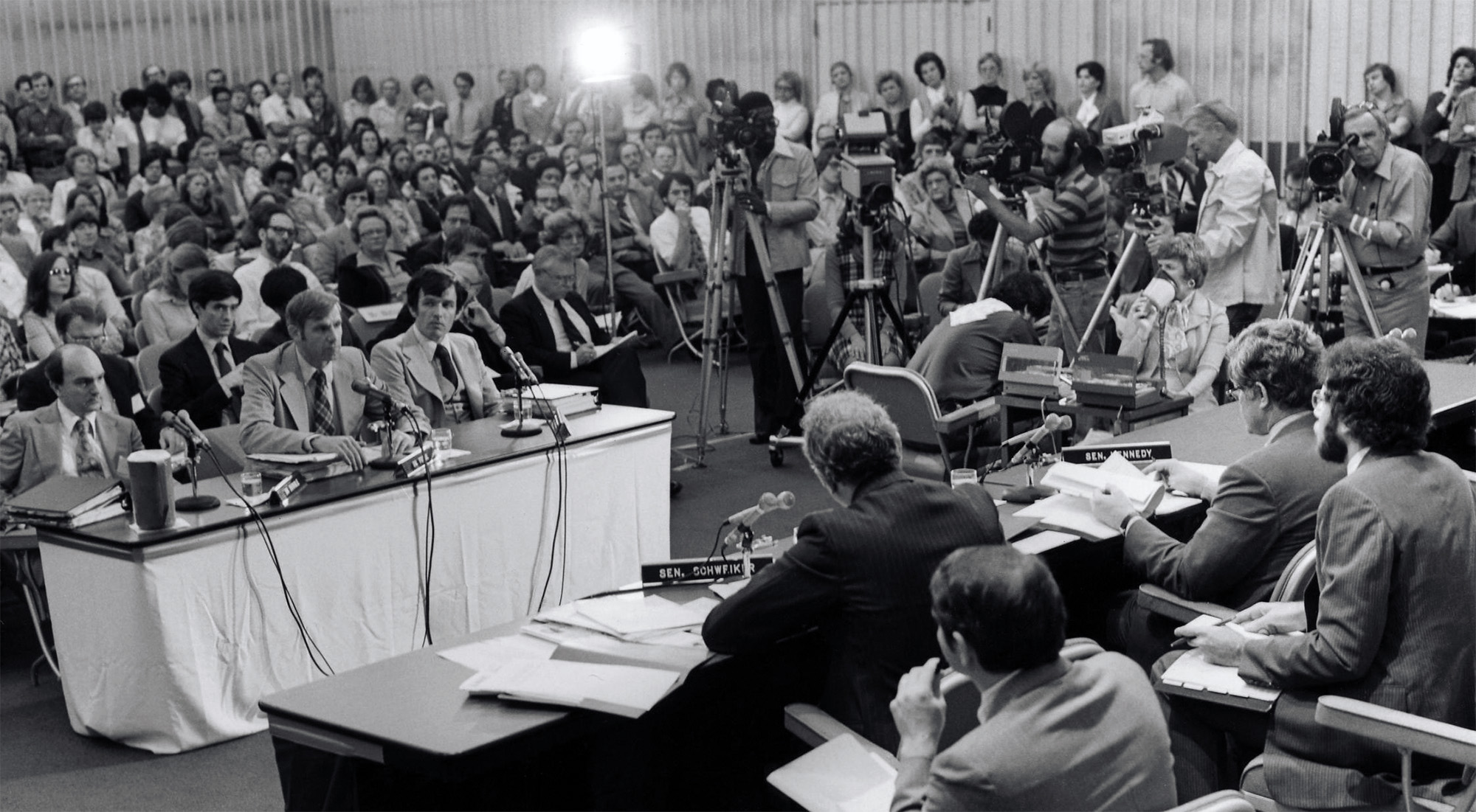 Men seated at two tables facing one another in a large room surrounded by a crowd. Several camera operators are visible in the background.