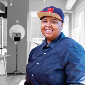 Tia Lyles-Williams, smiling in a sunny room, wearing blue button down shirt and blue baseball hat