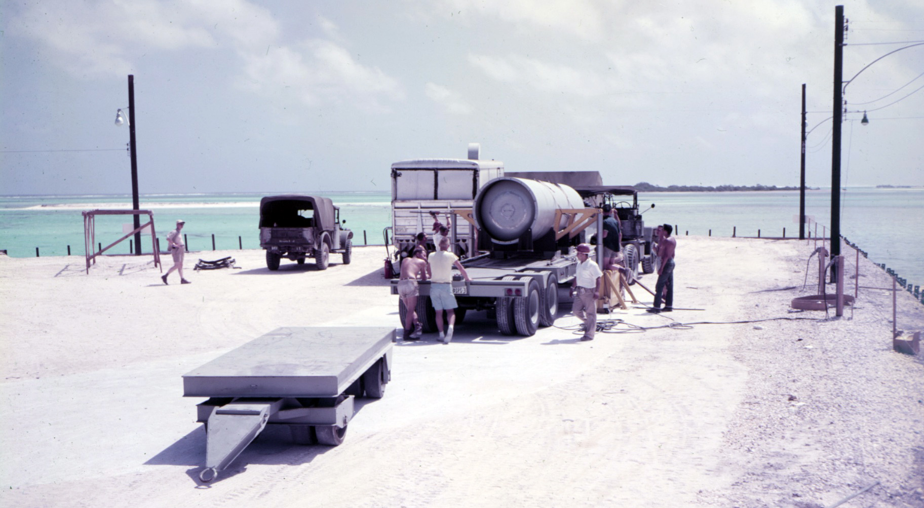 A group of men standing around a truck on a beach. A large cylindrical device is on the bed of the truck.