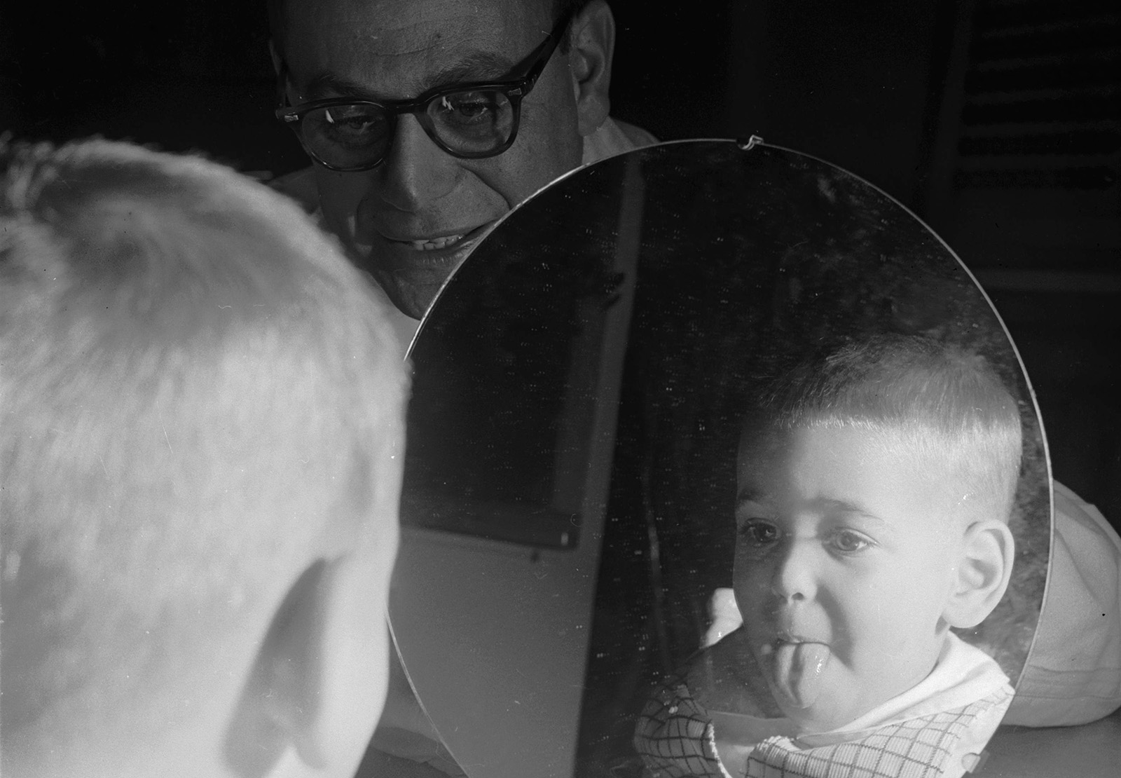 An infant sticks its tongue out and observes its reflection as a man looks on from behind the mirror