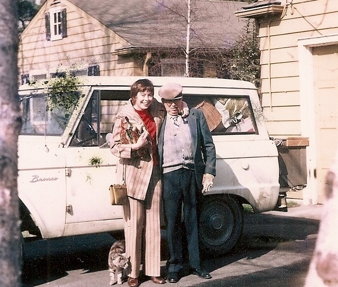 a man and woman standing in front of a house and car with a cat