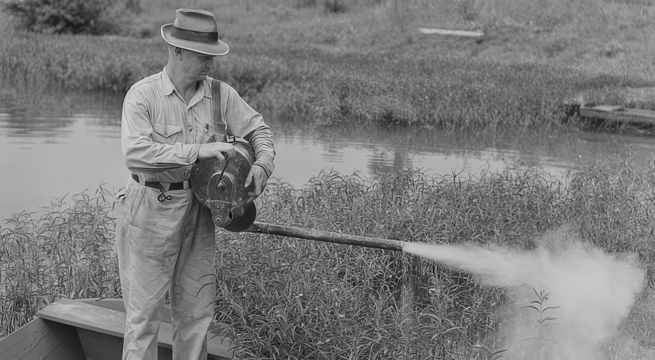 A man standing on a boat holding a spraying device that is emitting a cloud of white gas in a marshy environment