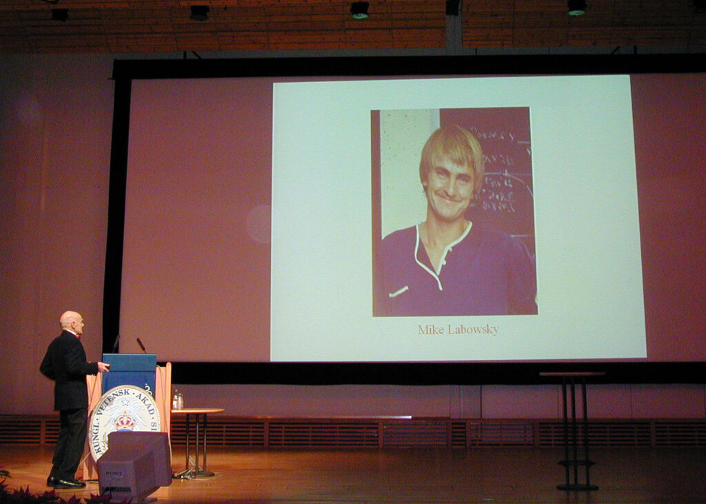 A man at a podium on a stage looking up at a projector screen. The slide on the screen shows a photograph of a smiling man with the caption: “Mike Labowsky.”