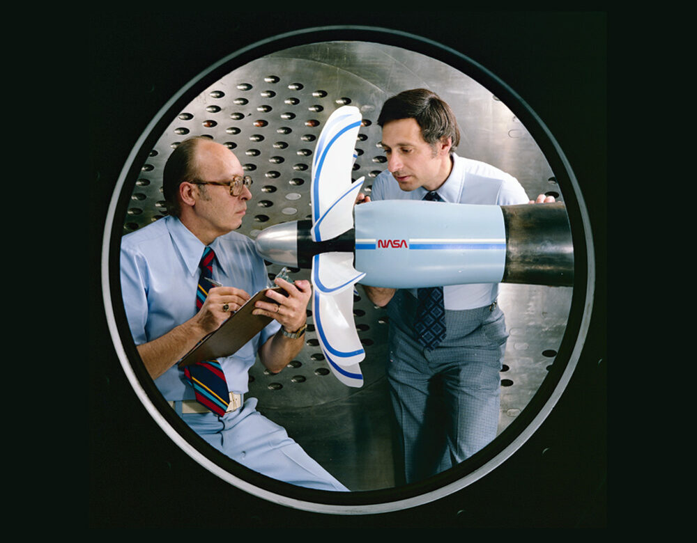 Two men standing next to a plane propeller in a room with metal walls.