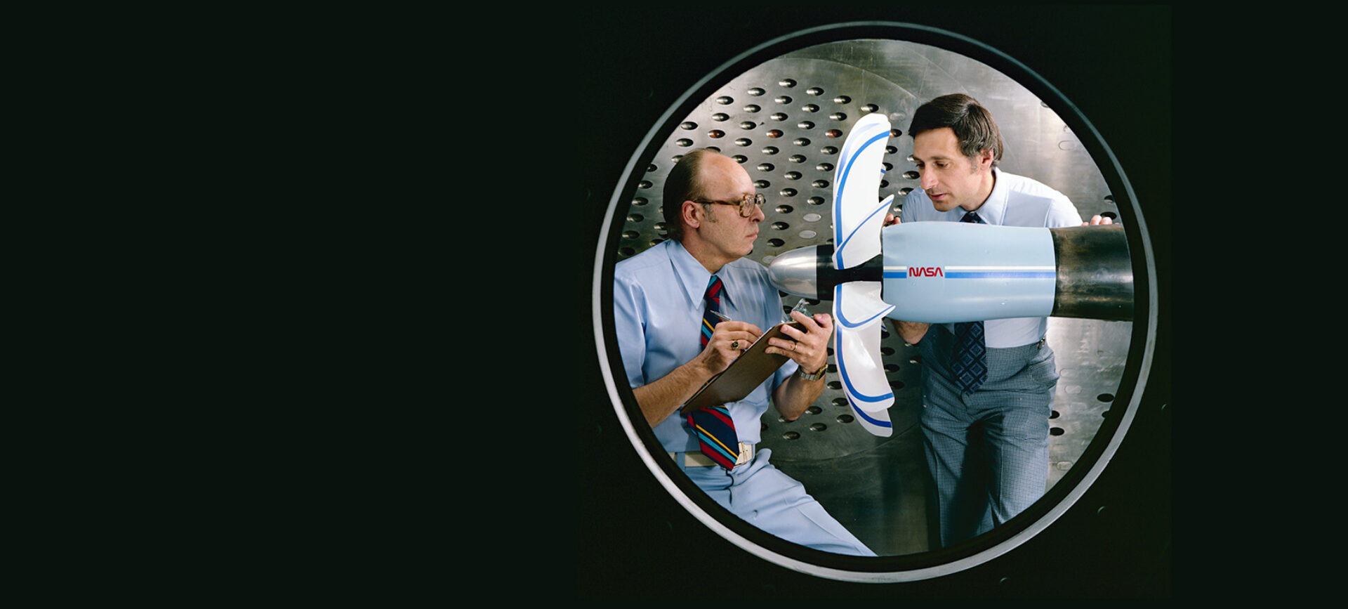 Two men standing next to a plane propeller in a room with metal walls.