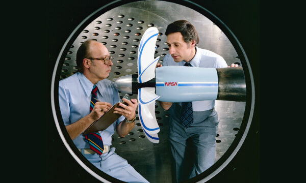 Two men standing next to a plane propeller in a room with metal walls.
